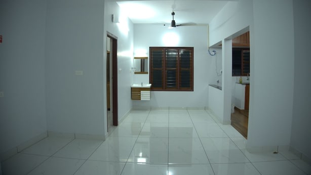 A spacious, well-lit hallway with a tiled floor, a ceiling fan, and a window with wooden shutters. - Pop Tavern Nakshatra Apartments  (Thiruvananthapuram)