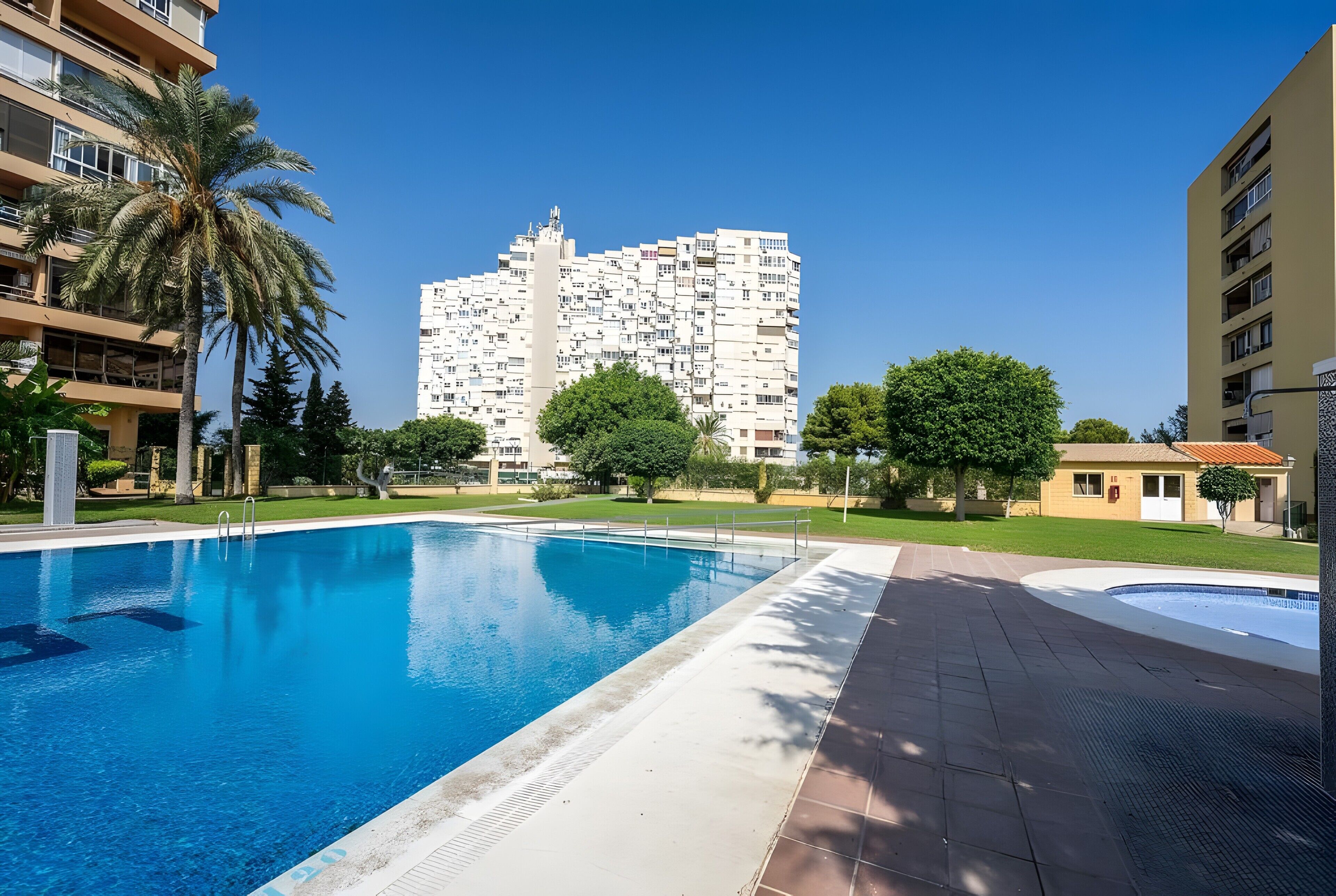 Une piscine à l’eau d’un bleu limpide, entourée d’une allée pavée. Au fond, on aperçoit un grand immeuble d’habitation doté de nombreux balcons.