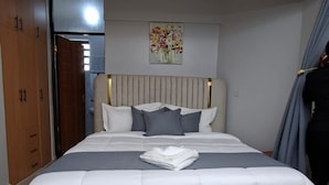 A bedroom with a large bed, a headboard, a wall-mounted light fixture, and a door leading to another room. - Novara Hotel Mwihoko  (Nairobi)