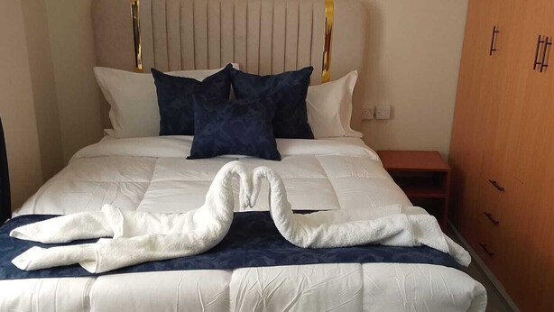A bed with a white duvet and two dark pillows, a wooden nightstand with a drawer, and a wooden wardrobe with a handle. - Novara Hotel Mwihoko  (Nairobi)