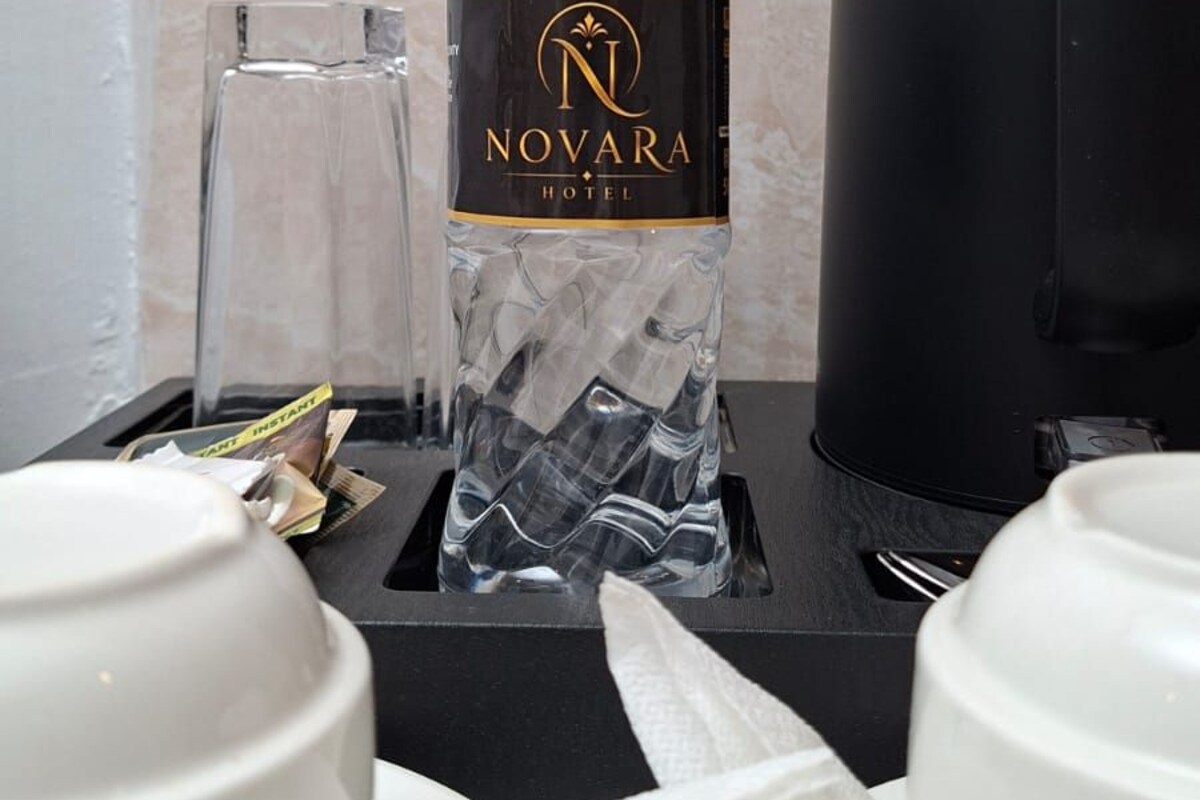 A minibar setup with a bottle labeled 'NOVARA HOTEL,' a glass, a coffee maker, and white cups and saucers.