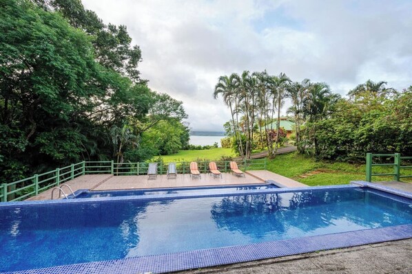 A swimming pool with a view of the sea, surrounded by greenery and a few lounge chairs.