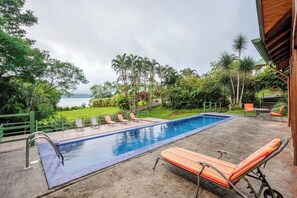 En pool med solstolar, utsikt över havet och frodig grönska.