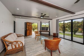 A spacious living area with a sofa, chairs, a glass-top coffee table, a television, and a sliding glass door leading to a garden.