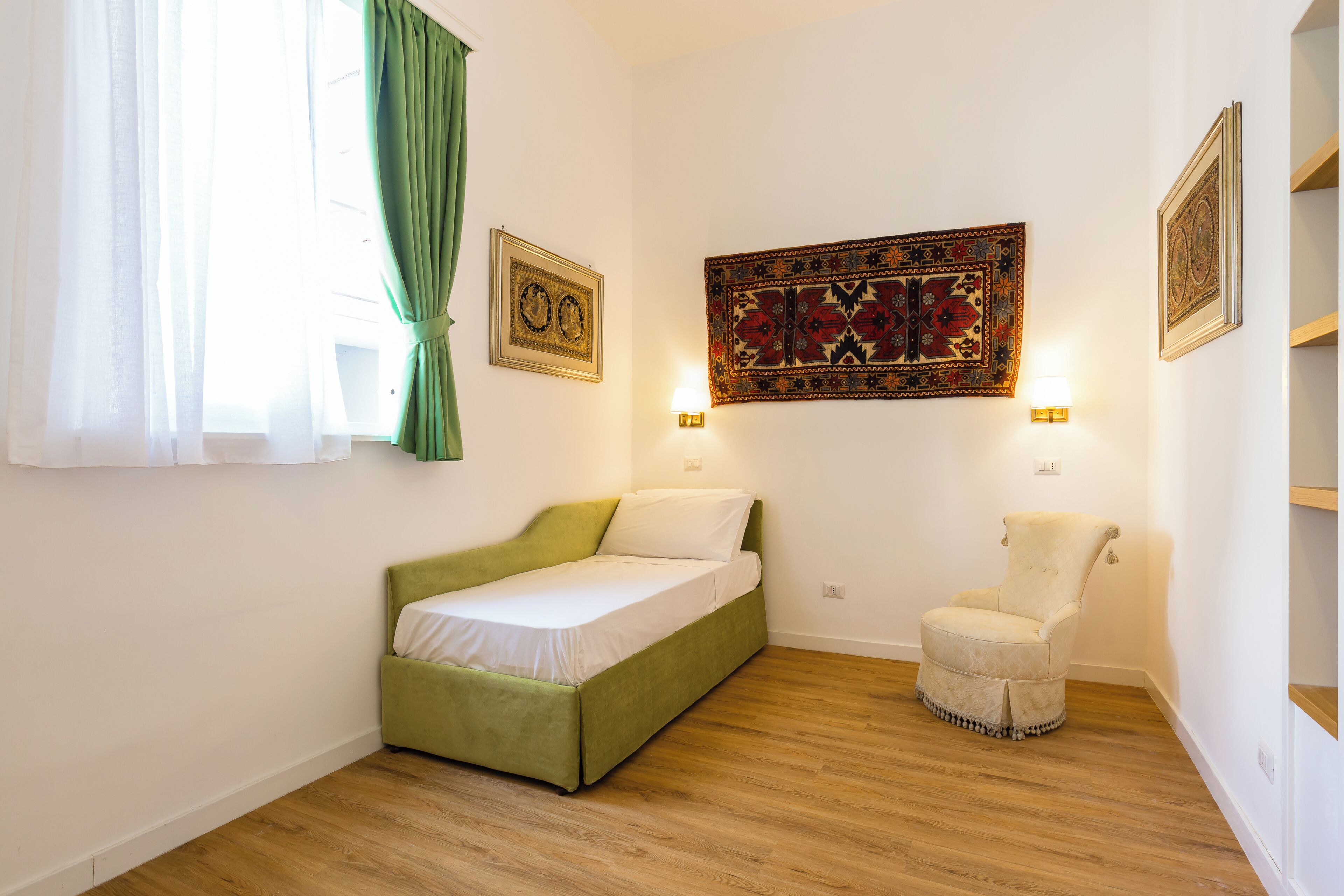 A small, neatly arranged room with a single bed, a chair, and a wall-mounted lamp.
