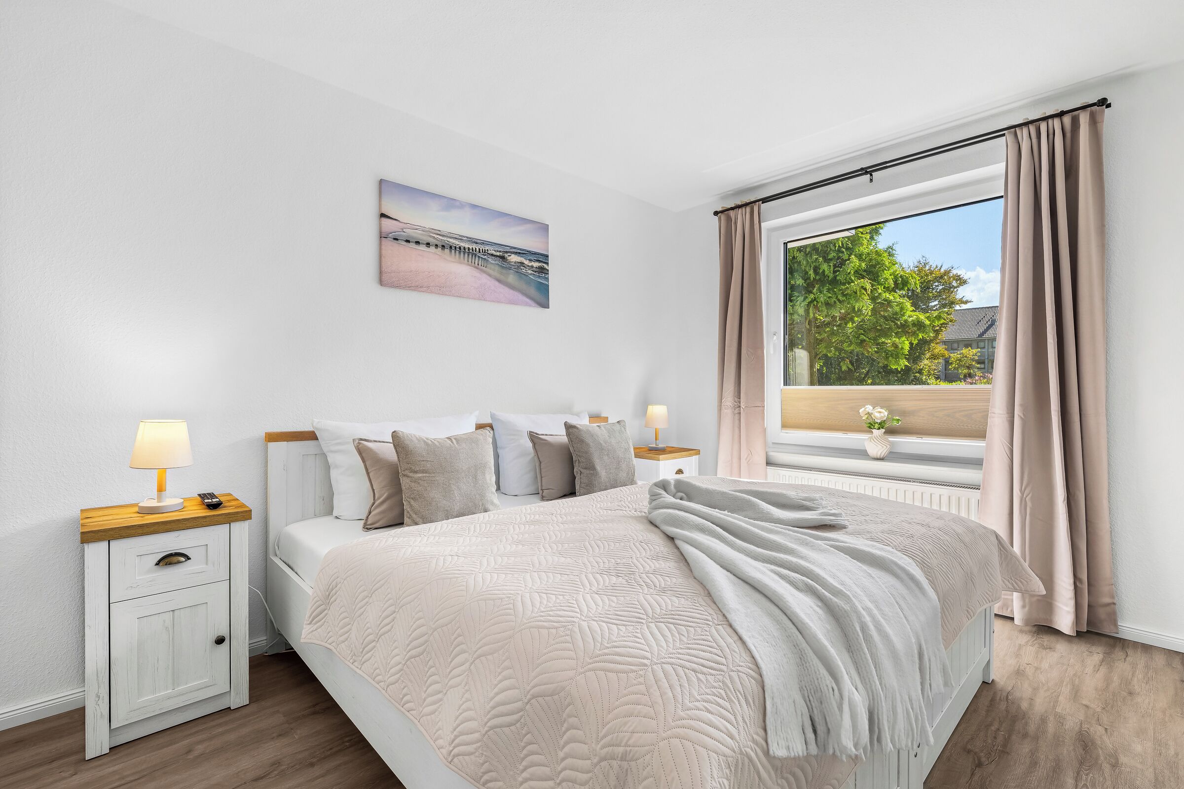 Un dormitorio con cama, mesitas de noche, un cuadro y una ventana con cortinas.