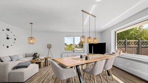 A modern living room with a sofa, coffee table, and dining table with chairs. There is a large window with a view of greenery and a door leading outside. - Blauort Vacation Home in Büsum: Spacious Luxury by the Sea for the Whole Family (Büsum)