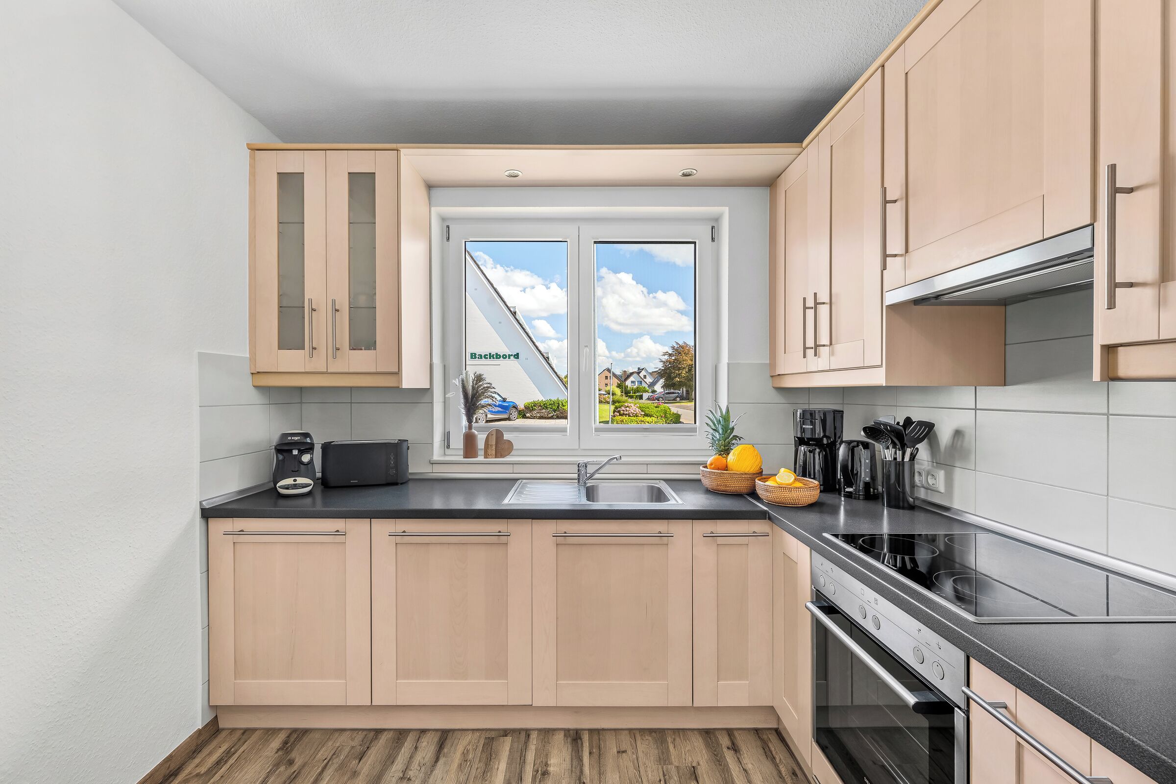 A modern kitchen with light wood cabinets, a black countertop, a stainless steel sink, and a view of a residential area through a large window.