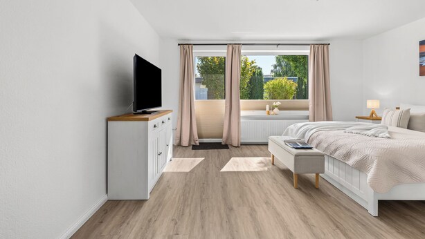 A modern bedroom with a flat-screen TV, a bed with a white duvet, a bench, and a window with curtains. - Blauort Vacation Home in Büsum: Spacious Luxury by the Sea for the Whole Family (Büsum)