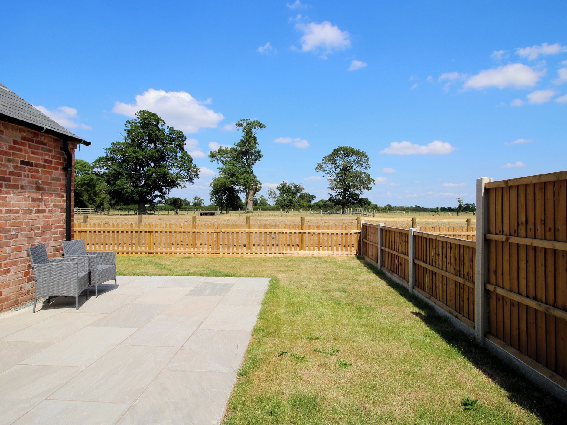 A patio area with a brick building, outdoor furniture, a wooden fence, and a grassy field.