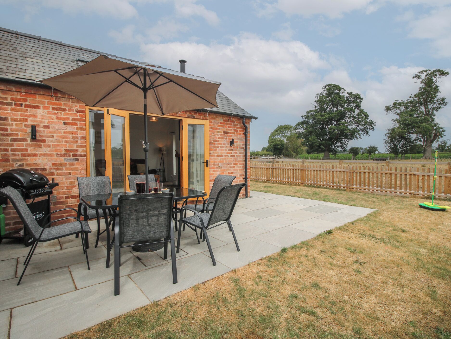 Un patio con una casa de ladrillo, mobiliario exterior, una parrilla y un parasol.