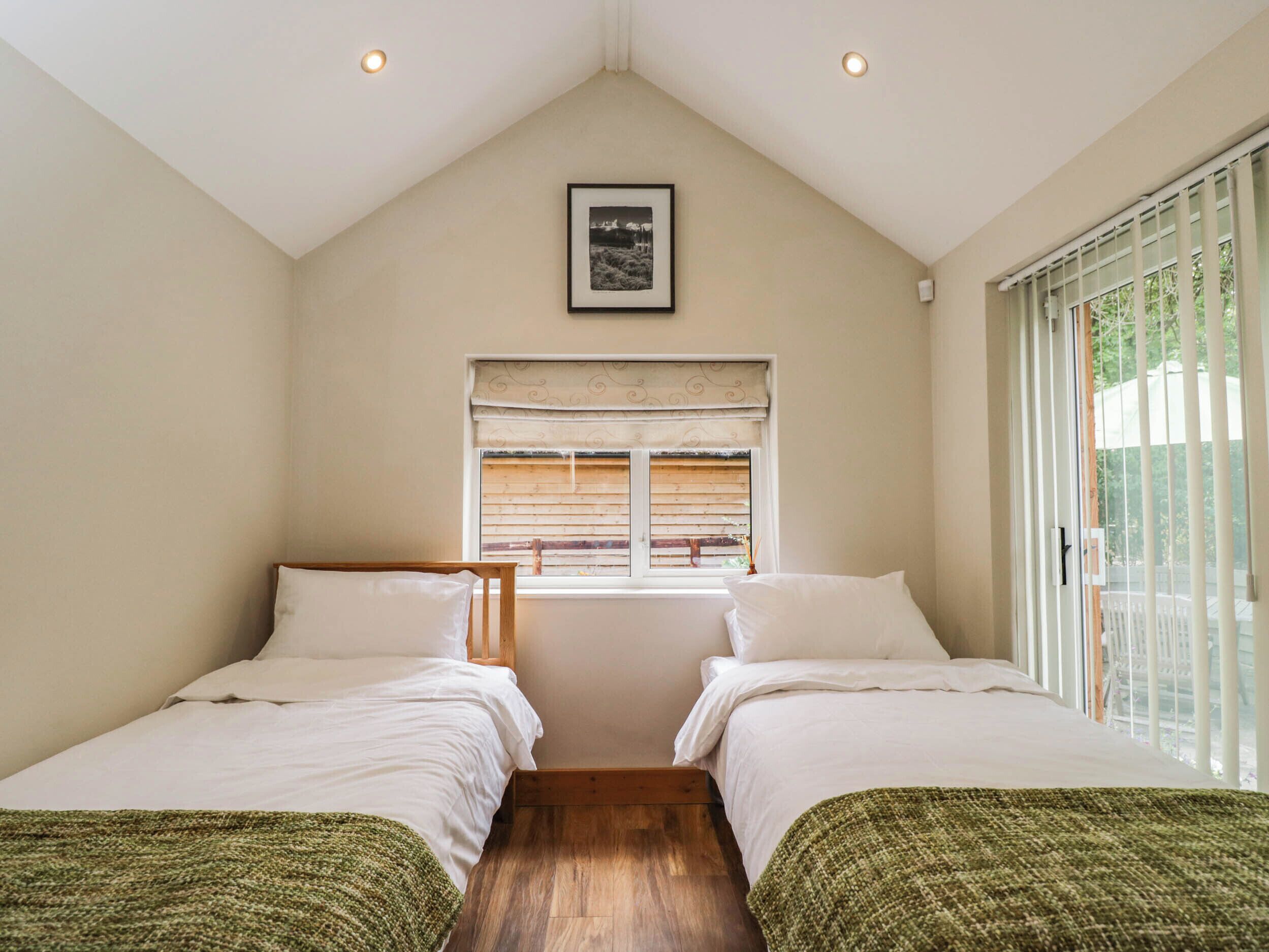 A bedroom with two beds, a window with blinds, a ceiling light, and a framed picture on the wall.