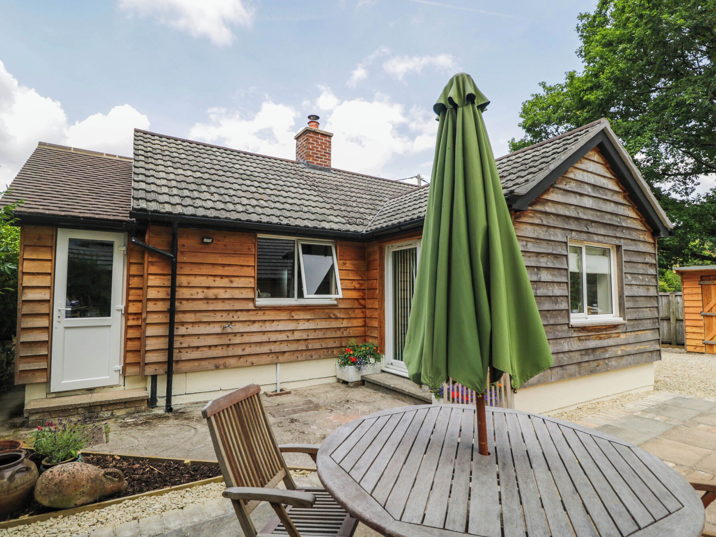 A wooden house with a tiled roof, a patio table with a green umbrella, and a wooden chair.