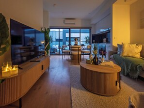 Ein modernes Wohnzimmer mit Flachbildfernseher, Couchtisch und Sofa. Es gibt einen Essbereich mit Stühlen und Tisch sowie eine Küchenzeile mit Schränken und Arbeitsplatte.