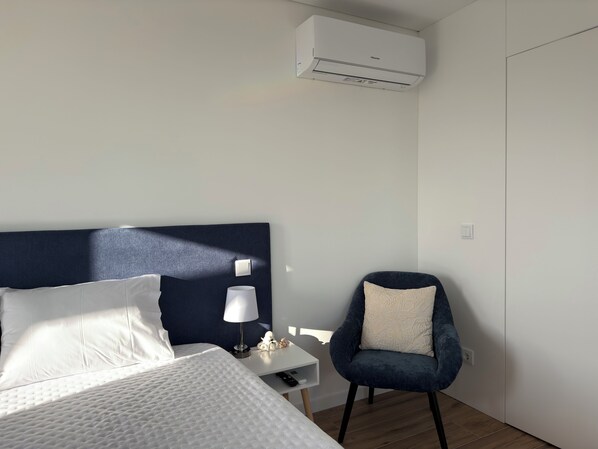 A hotel room with a bed, a chair, a nightstand, a lamp, and an air conditioning unit.