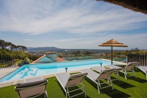 Ein Swimmingpool mit Blick auf die Landschaft, umgeben von Liegestühlen und einem Reetdachschirm.