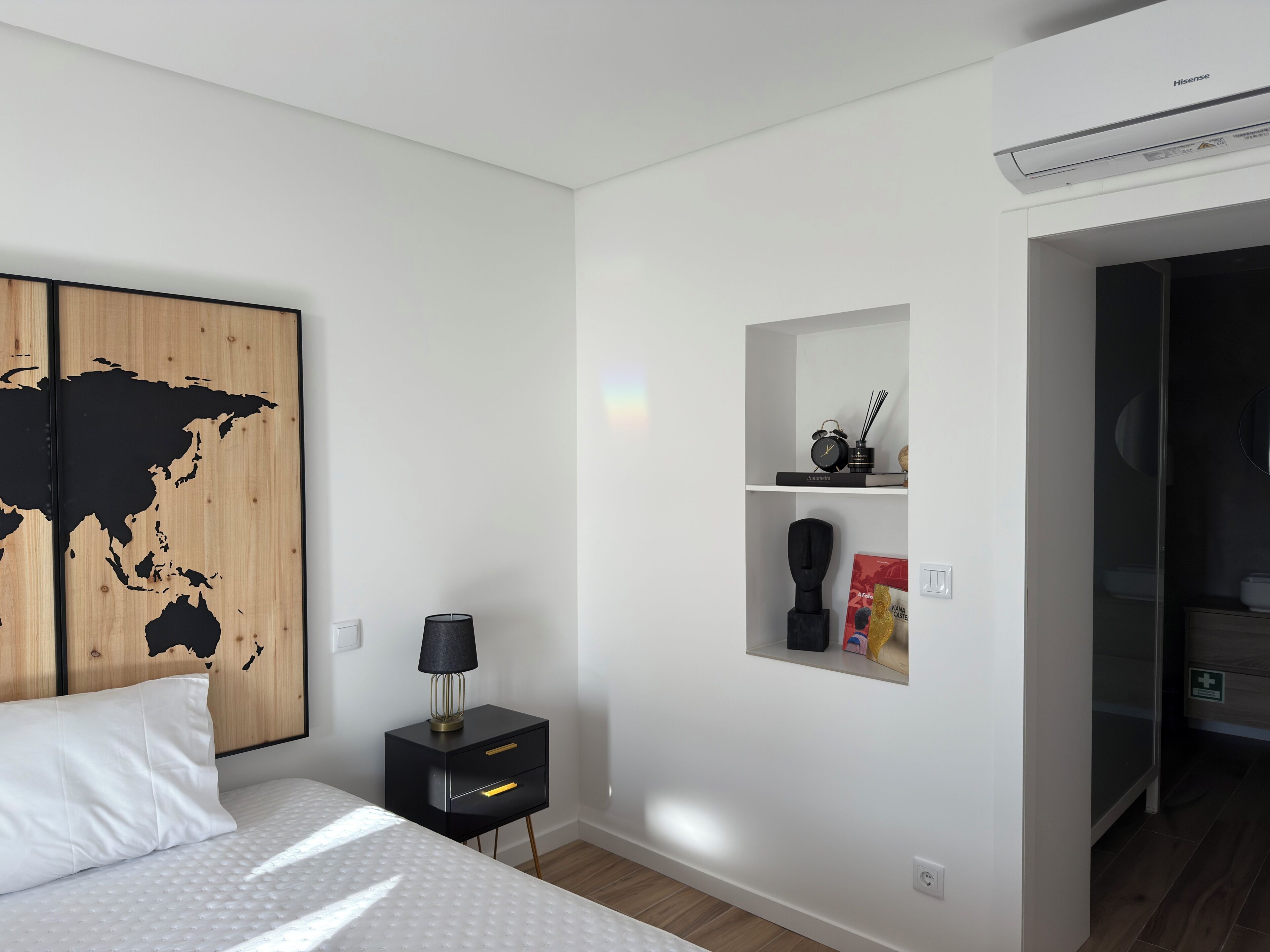 A modern bedroom with a bed, a nightstand, a world map artwork, a shelf with decorative items, and a door leading to a bathroom.