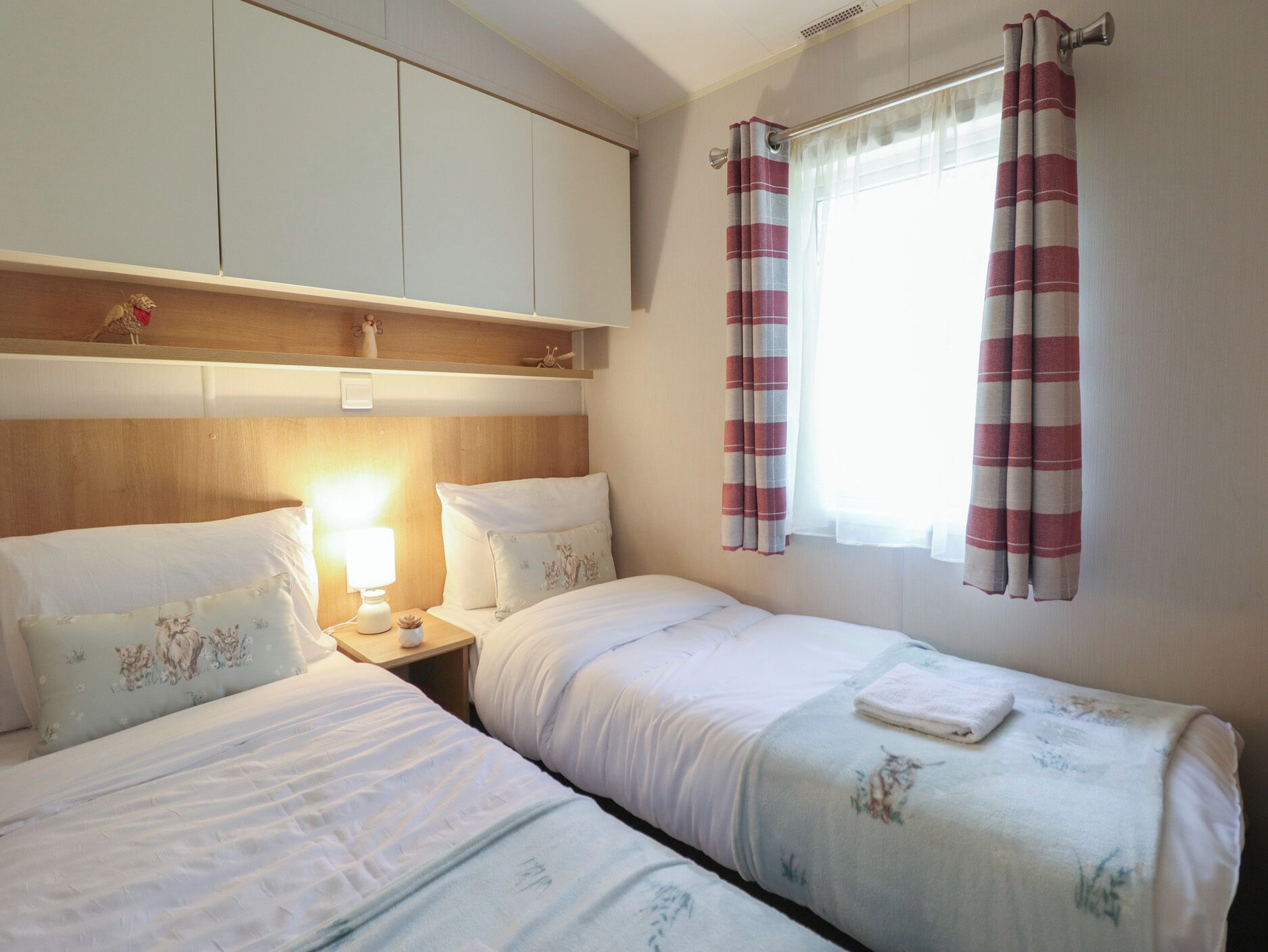Quarto de hotel com duas camas, uma mesa de cabeceira, um abajur, uma janela com cortinas e uma cabeceira de madeira.