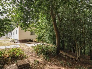 A mobile home with a small porch, surrounded by dense trees and a wooden deck. - Plot 13A (Ruthin)