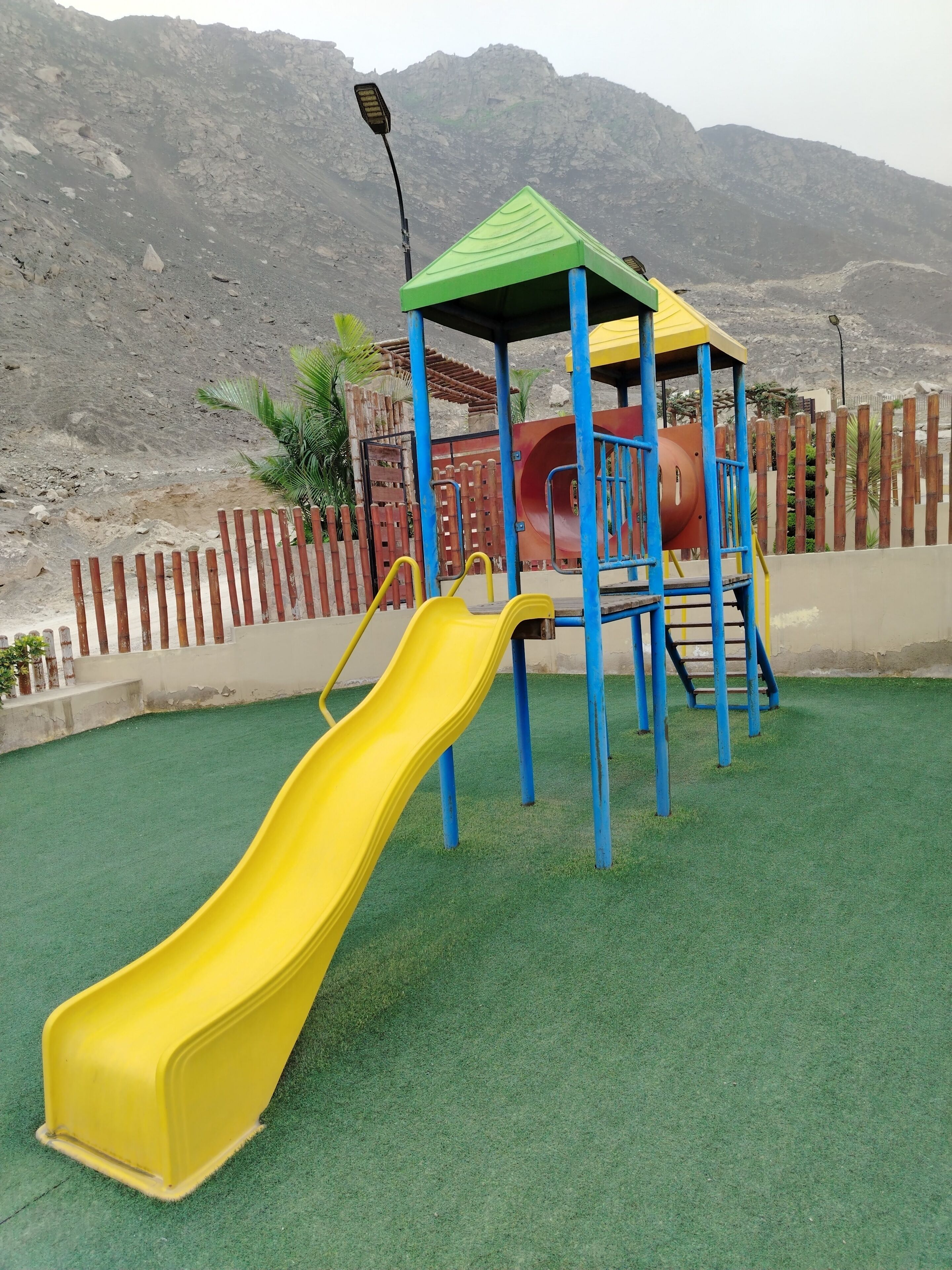 Un parque infantil con un tobogán amarillo y una estructura para escalar, rodeado por una valla de madera y un paisaje de montañas al fondo.