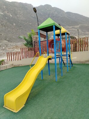 Ein Spielplatz mit einer gelben Rutsche und einer Kletteranlage, umgeben von einem Holenzaun und einem Berg im Hintergrund.