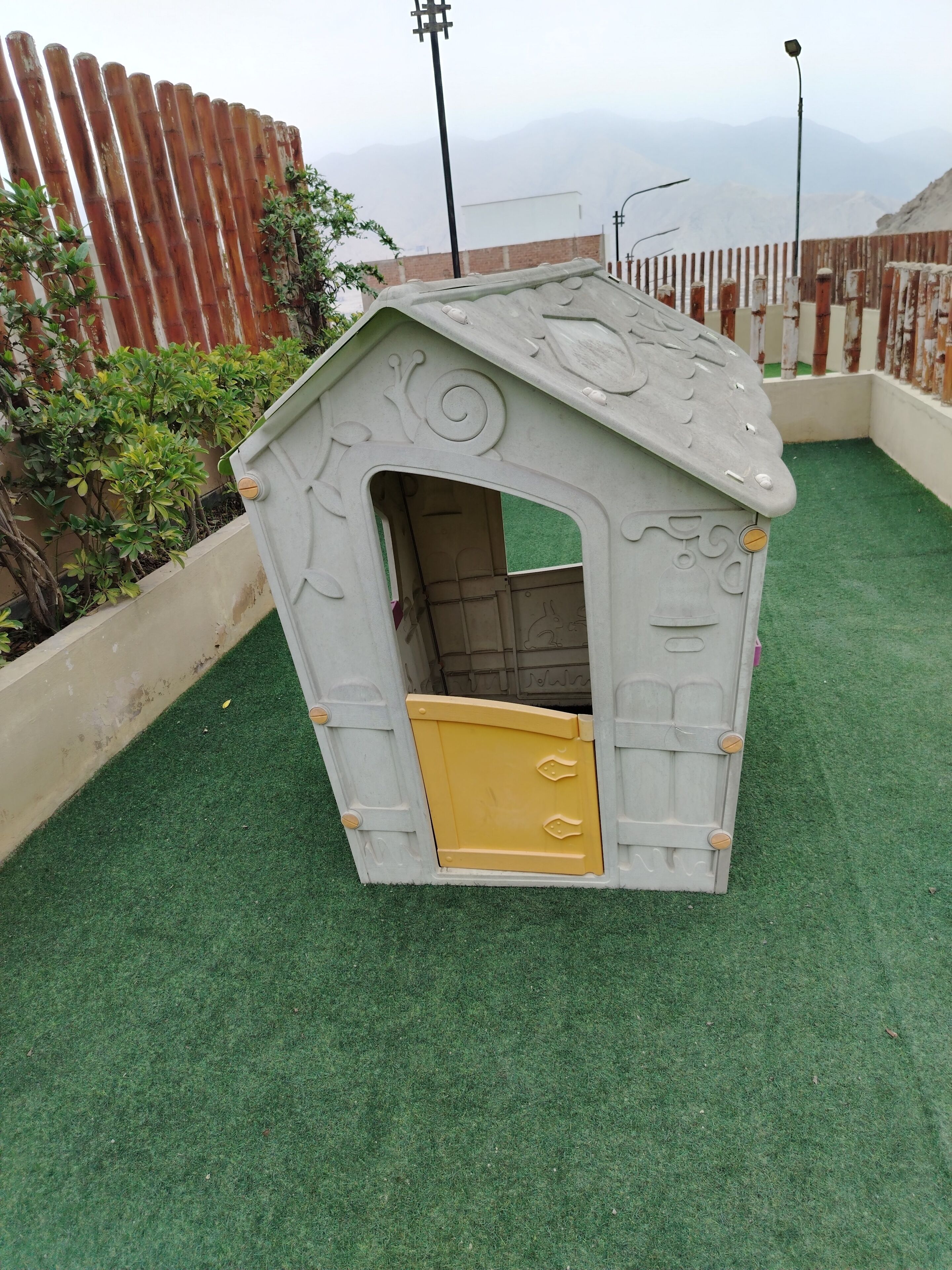 A small, enclosed playhouse with a door and a window, surrounded by a green artificial turf and a wooden fence.