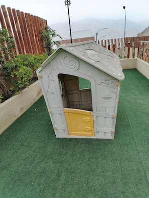 A small, enclosed playhouse with a door and a window, surrounded by a green artificial turf and a wooden fence.