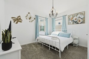 Une chambre à coucher avec un lit en métal blanc, une table de chevet, une commode, un lustre suspendu et un tableau accroché au mur.