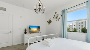 Une chambre à coucher avec un lit, un téléviseur posé sur une commode, un lustre et une fenêtre donnant sur des immeubles.