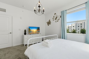Ein Schlafzimmer mit einem Bett, einem Fernseher auf einer Kommode, einer Kristallleuchter und einem Fenster mit Blick auf Gebäude.