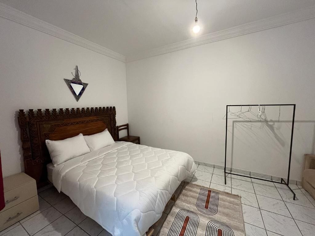 A bedroom with a wooden headboard, a bed with white bedding, a nightstand, a dresser, a wall-mounted light, and a radiator.