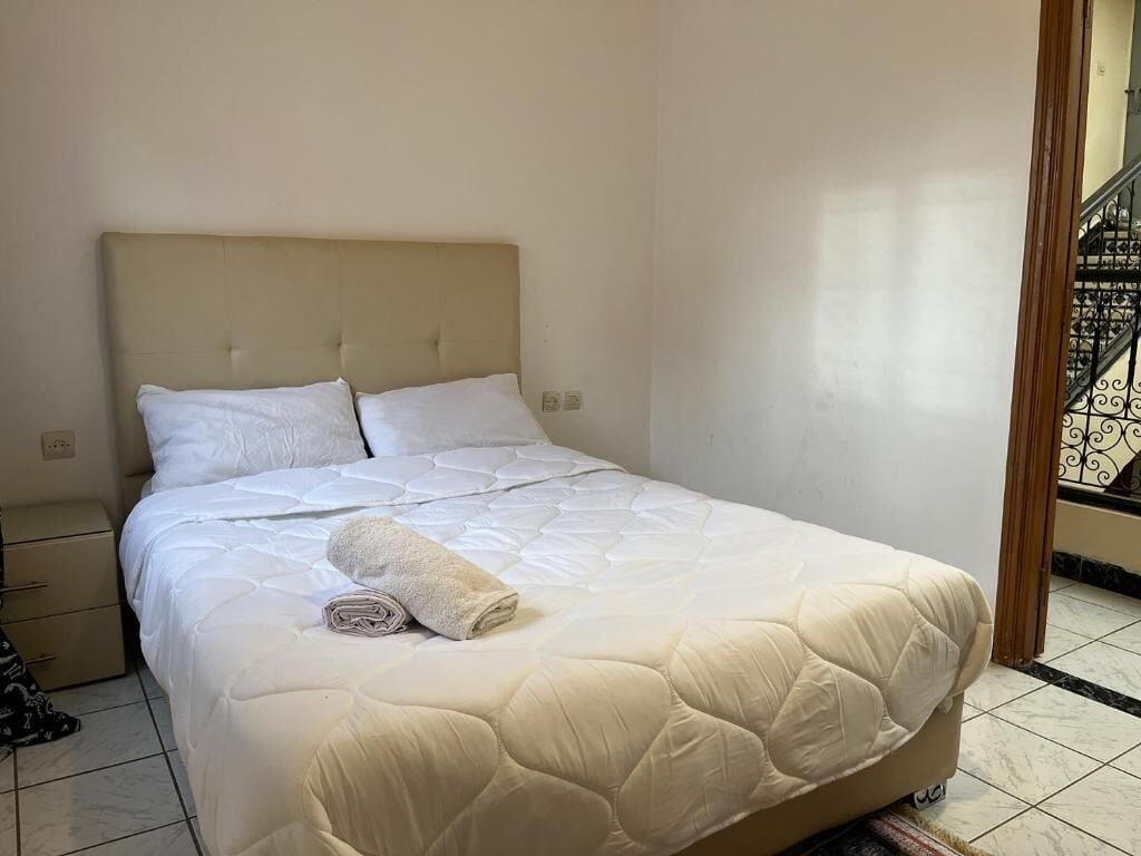 A neatly made bed with a quilted cover and a rolled towel, a bedside drawer, and a tiled floor.