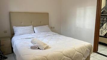 A neatly made bed with a quilted cover and a rolled towel, a bedside drawer, and a tiled floor.
