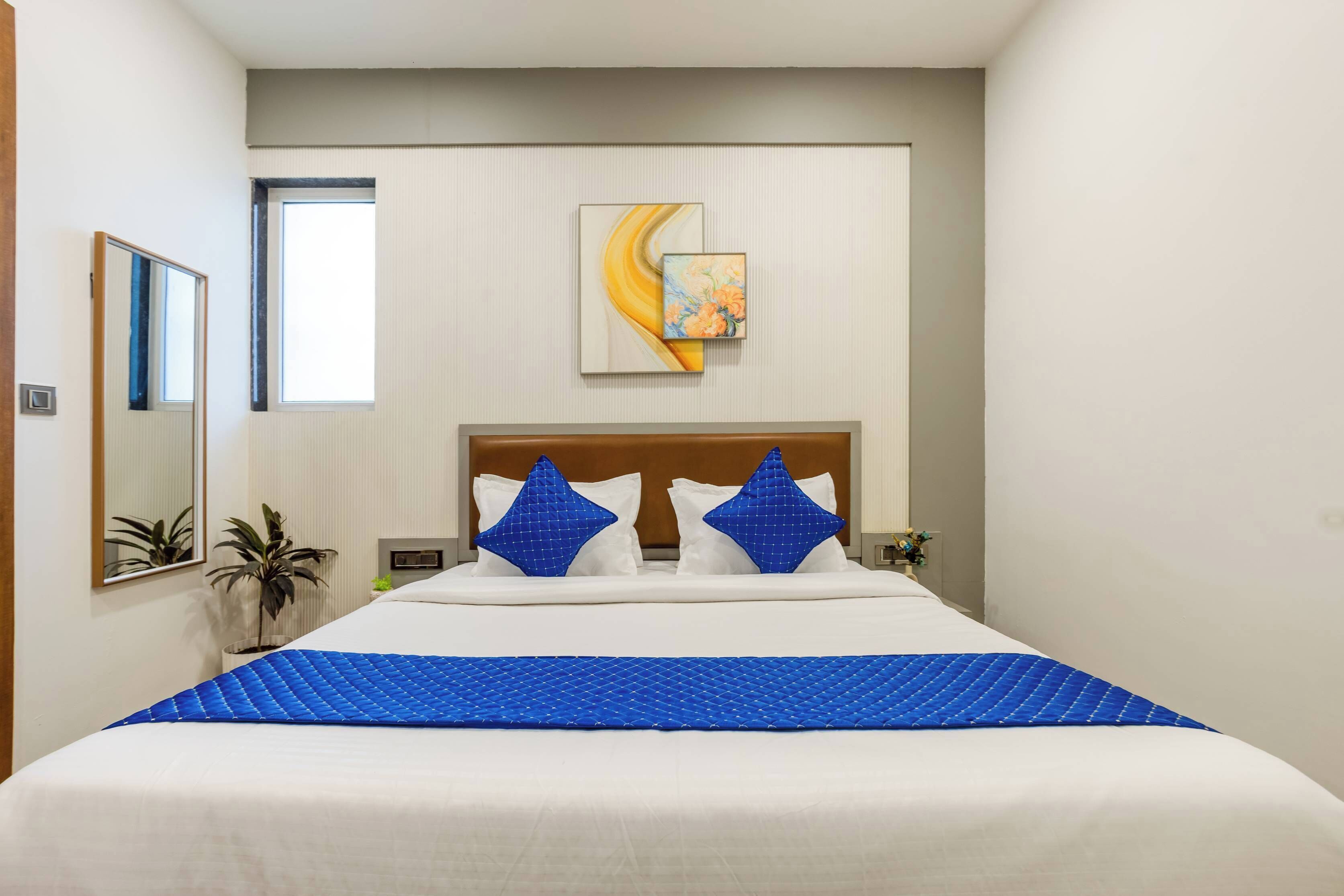 Une chambre d’hôtel avec un grand lit, deux oreillers bleus, un tableau au mur et une plante en pot.