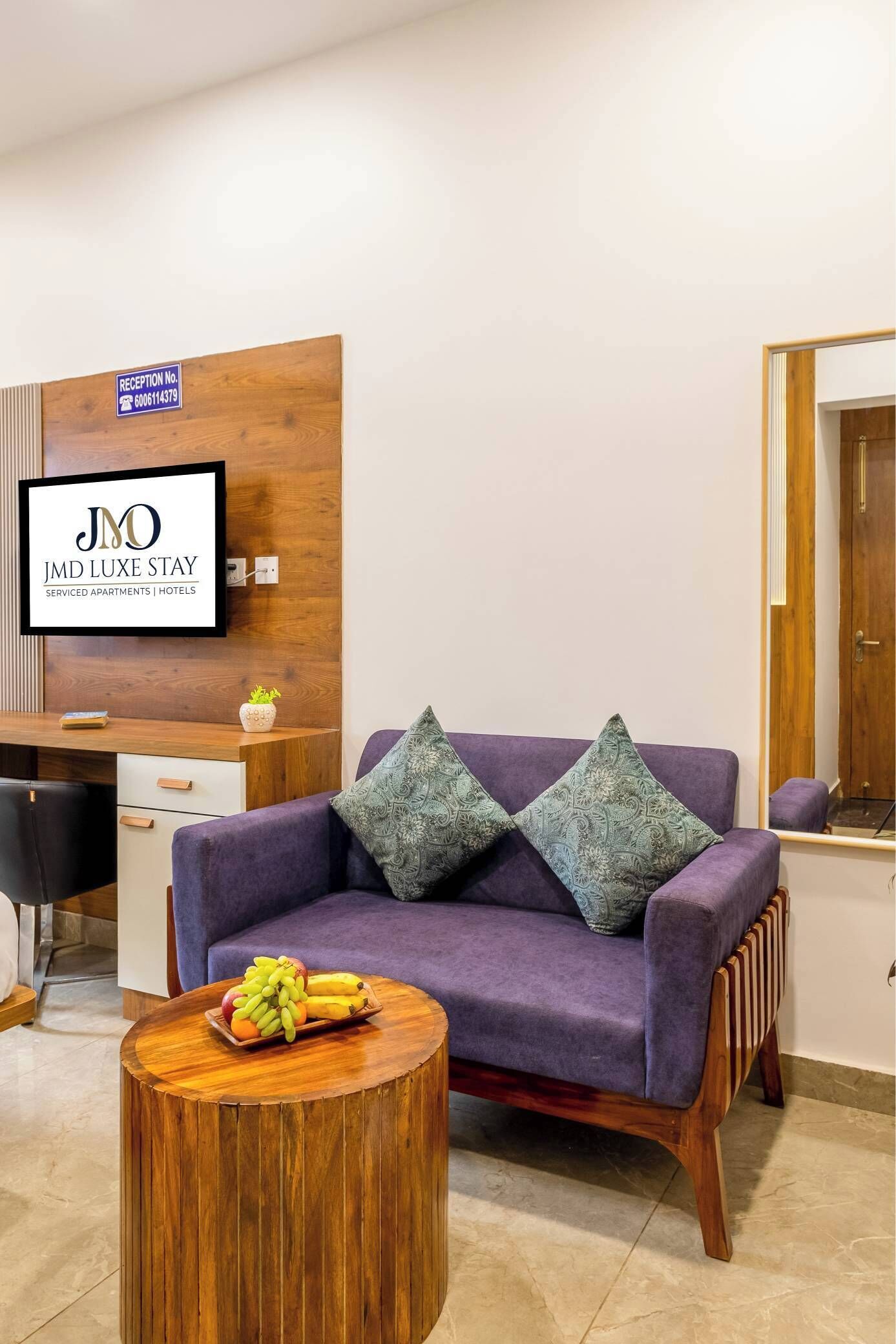 A reception area with a reception desk, a sofa with patterned cushions, a round wooden table with fruit, and a sign indicating serviced apartments and hotels.