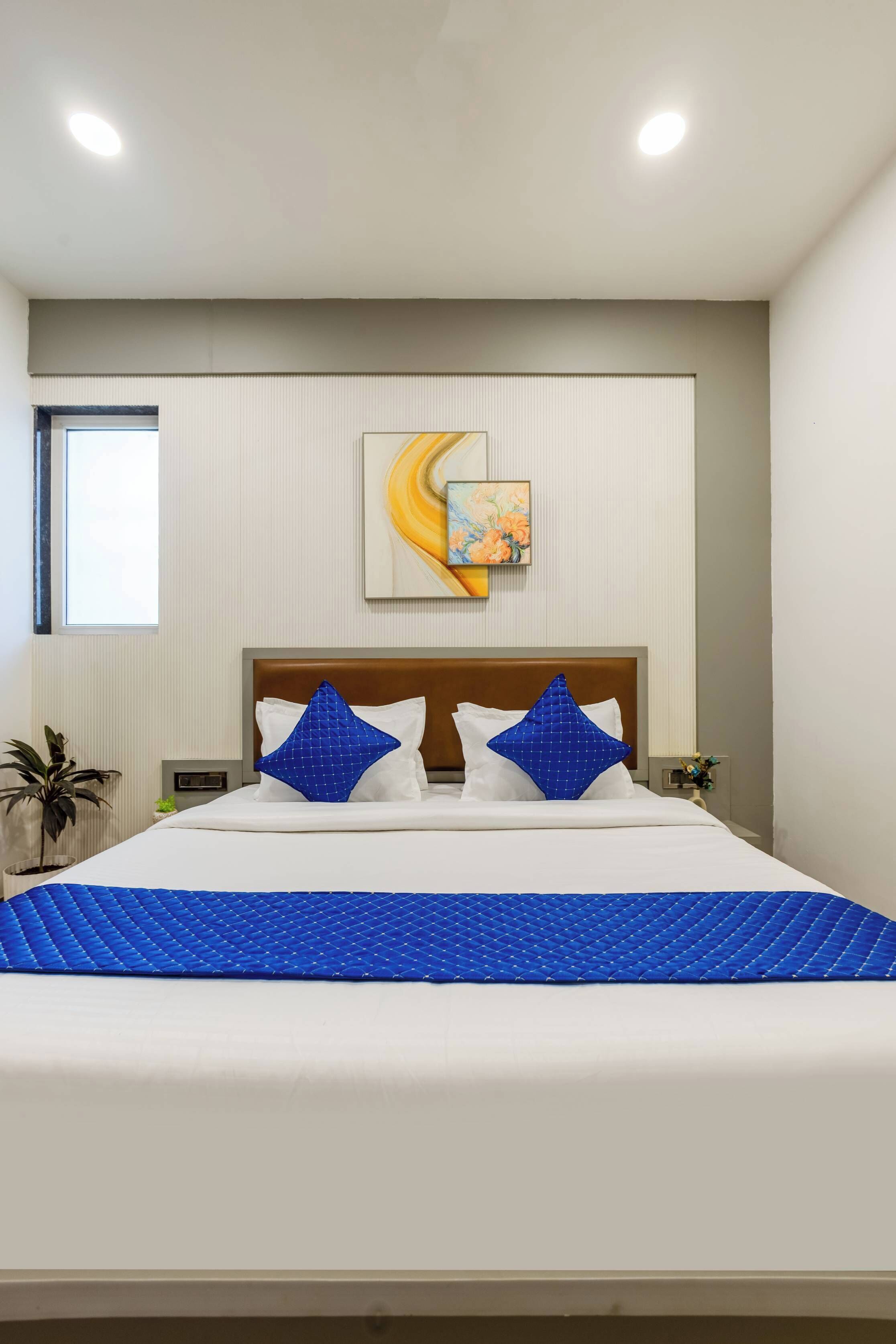 A hotel room with a bed, a window, a painting on the wall, and a plant.