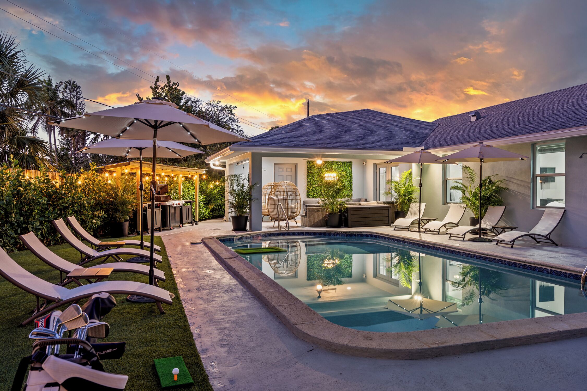 A swimming pool with lounge chairs and umbrellas, surrounded by a house with a covered patio and a view of the sky.