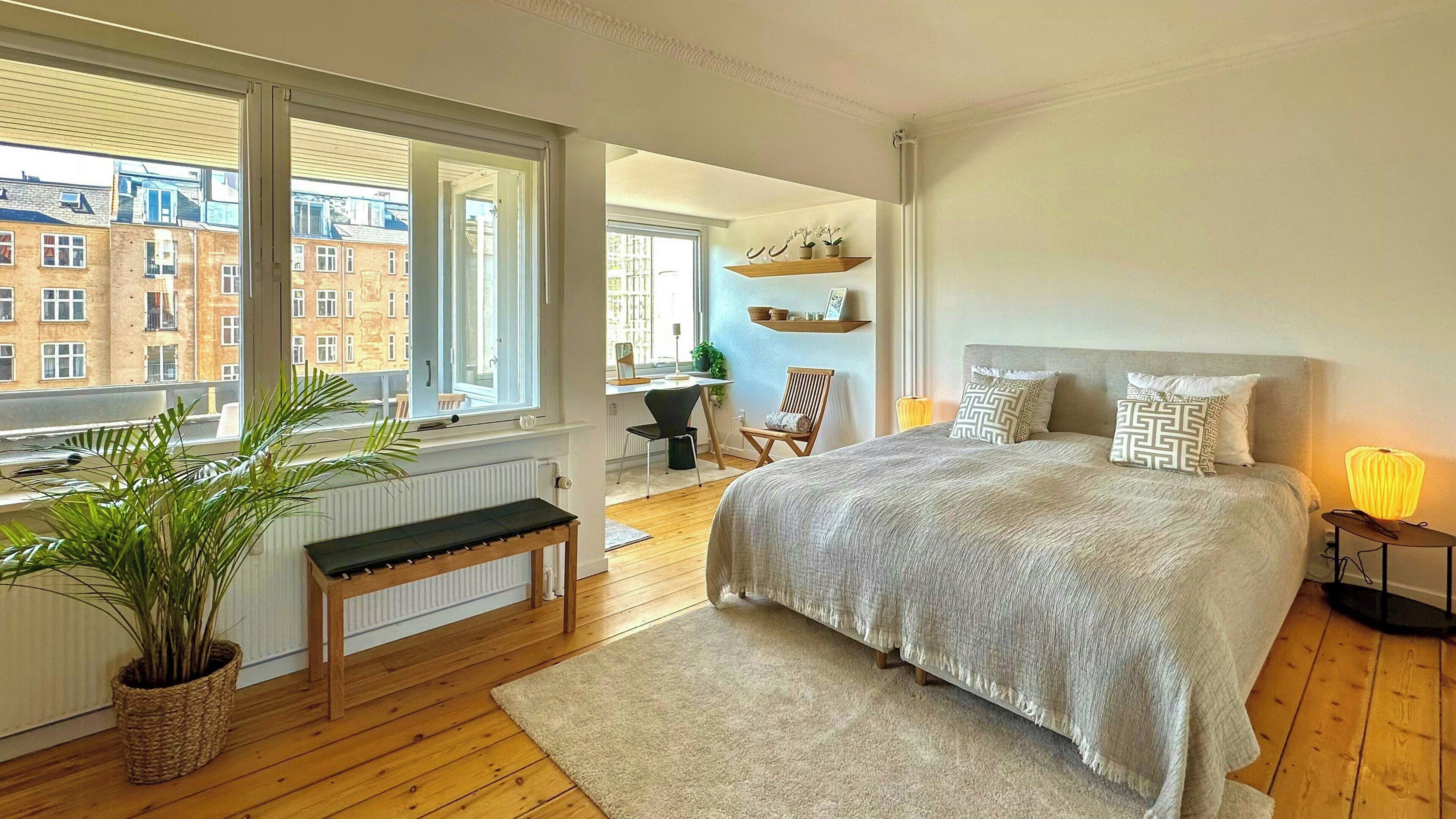 A bedroom with a large bed, a desk, and a view of buildings through the window.