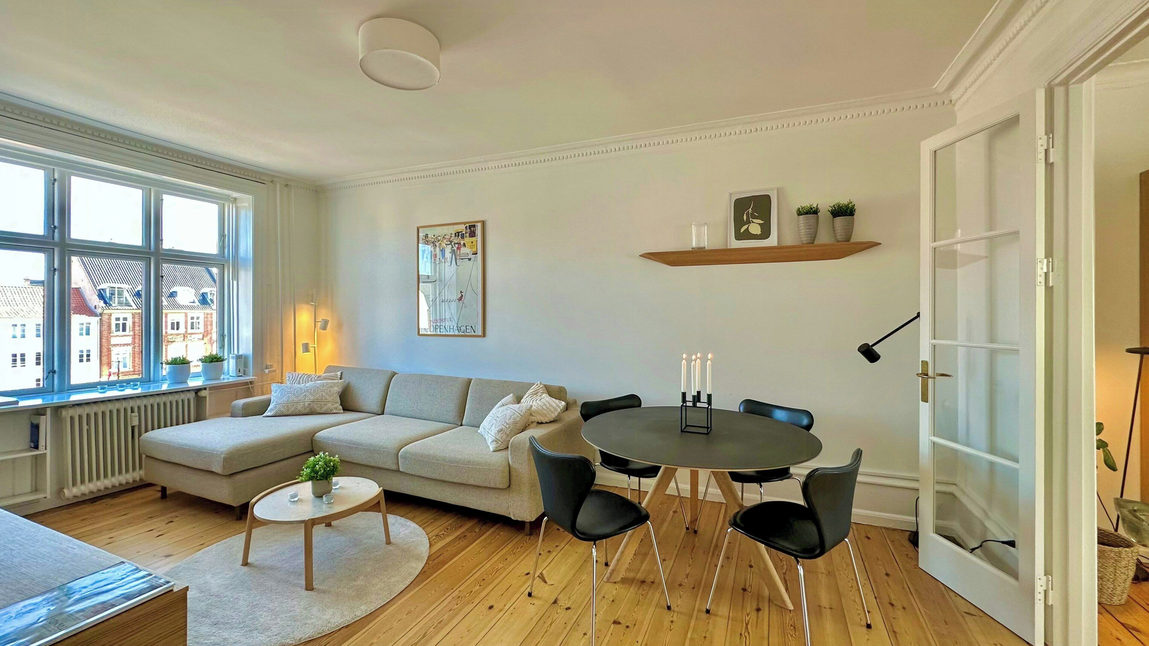 A modern living room with a sofa, round dining table, and chairs. There is a window with a view of buildings, a radiator, and a door.