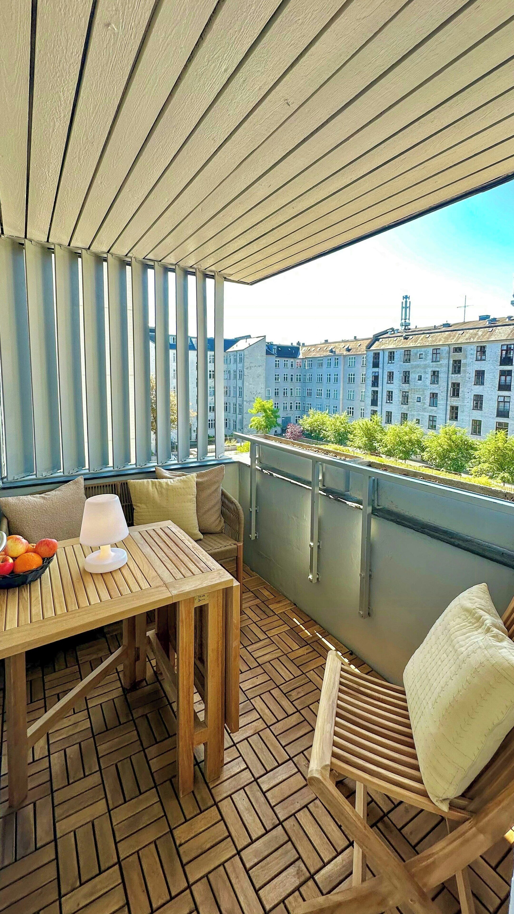A wooden deck with a table and chairs, a bowl of fruit, and a view of buildings in the distance.