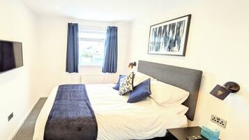 A hotel room with a bed, a bedside table, a TV, a window with curtains, and a framed picture on the wall.