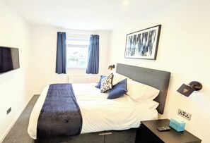 A hotel room with a bed, a bedside table, a TV, a window with curtains, and a framed picture on the wall. - The Kelburn - 4 bedroom house - Kilmarnock (Riccarton)