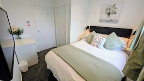 A hotel room with a bed, a bedside table with a vase of flowers, a wall-mounted light, and a door. - Kelburn, Kilmarnock - 4 bed house (Riccarton)