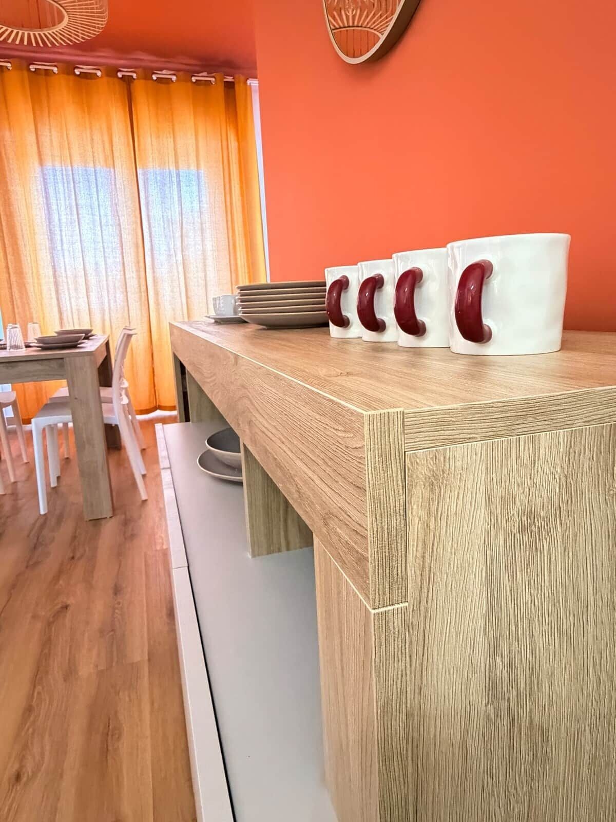Een houten aanrecht met witte mokken en een eettafel met stoelen.