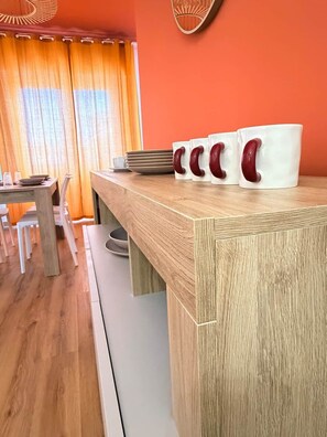 A wooden kitchen counter with white mugs and a dining table with chairs.