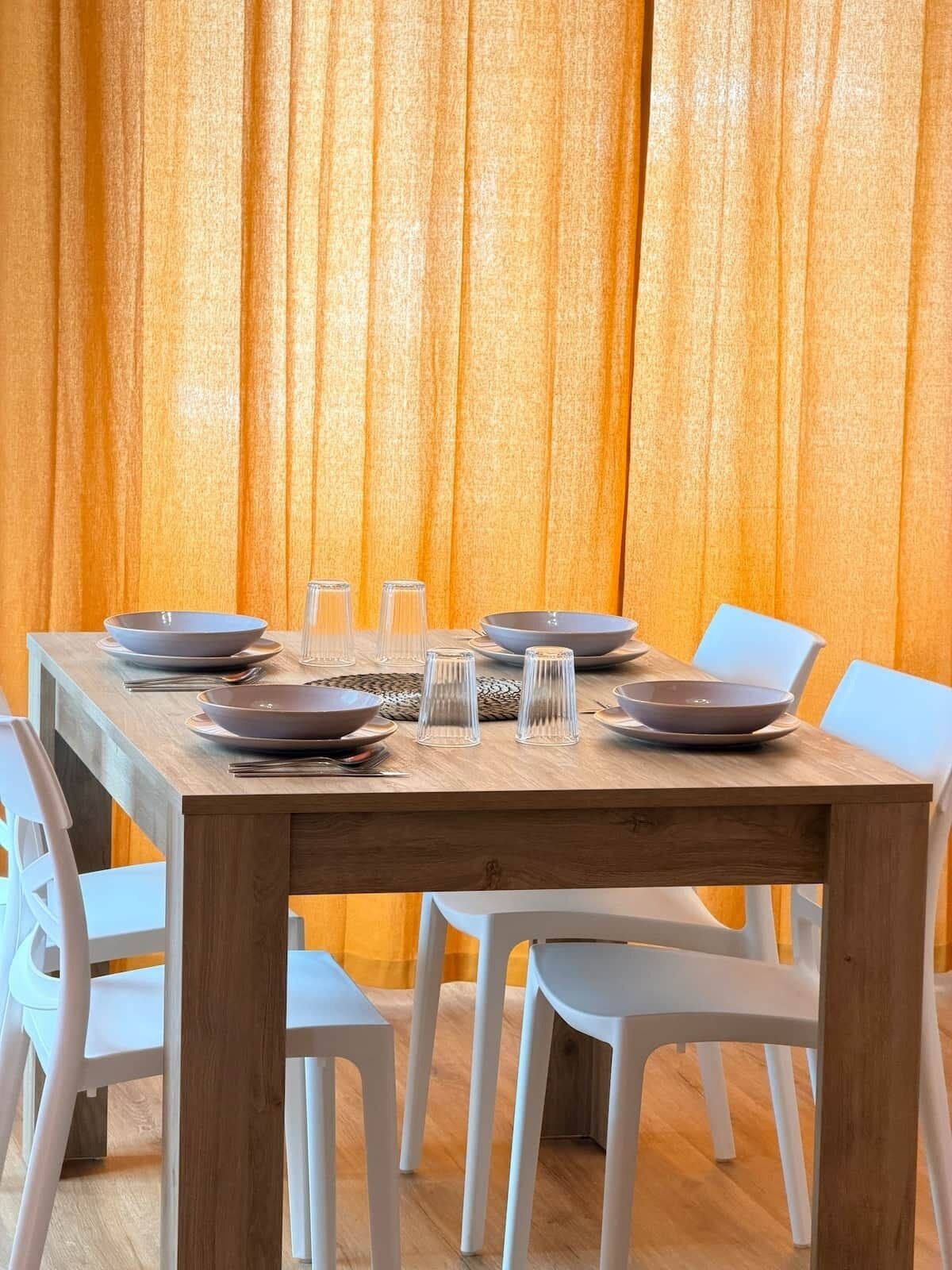 A dining table set with plates, bowls, and glasses, surrounded by white chairs, against a backdrop of orange curtains.