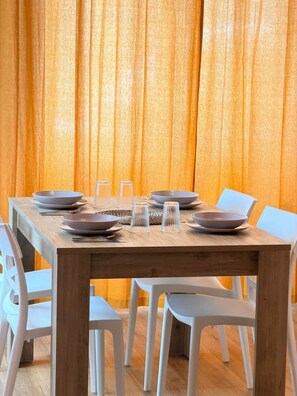 A dining table set with plates, bowls, and glasses, surrounded by white chairs, against a backdrop of orange curtains.