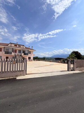 A two-story residential building with a gated entrance, a paved driveway, and a view of the surrounding landscape.