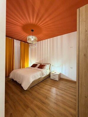 A bedroom with a wooden floor, a bed with white bedding and brown pillows, a bedside table with a lamp, and a ceiling light fixture.