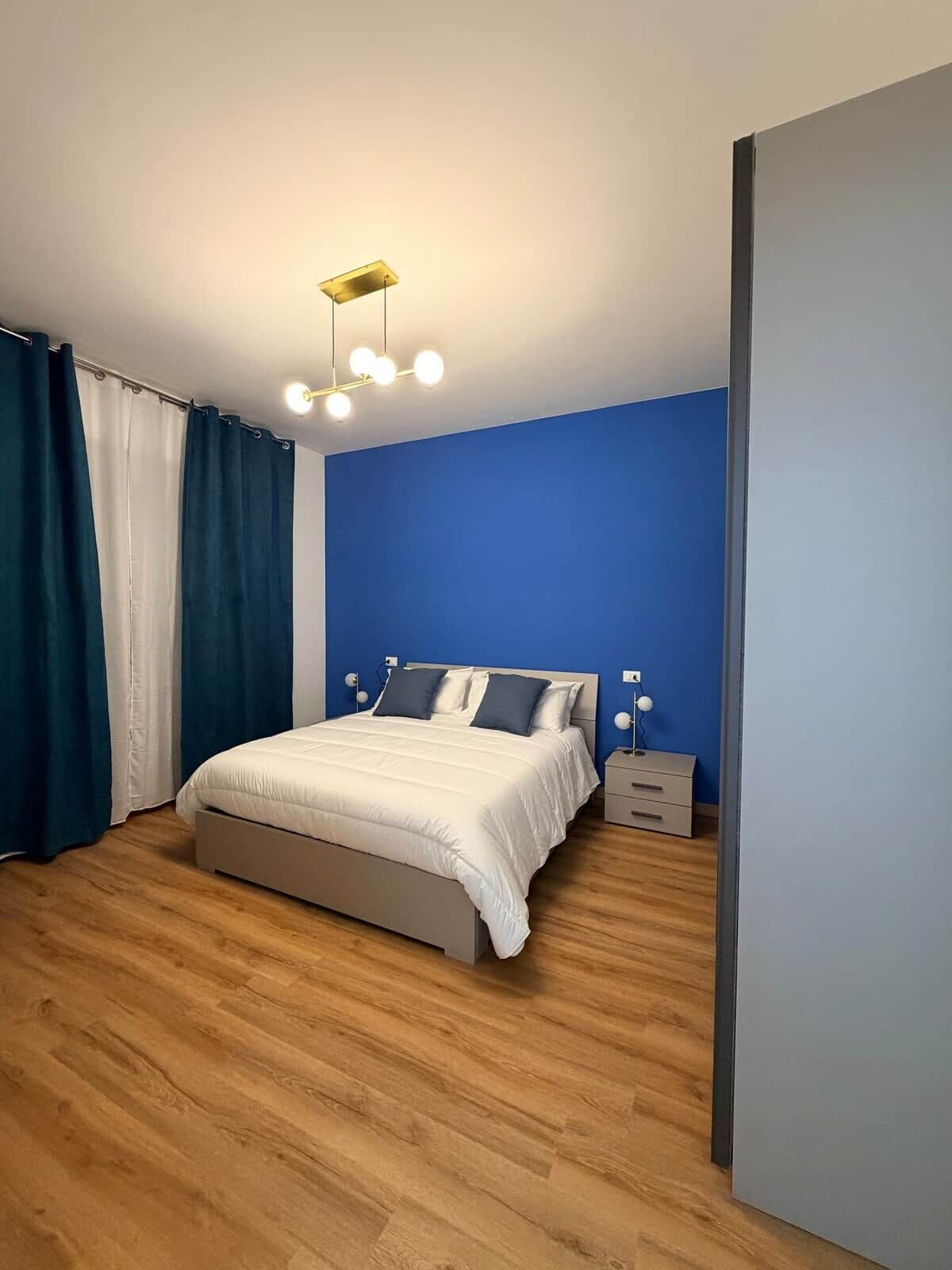 A bedroom with a bed, curtains, a nightstand, and a wall-mounted light fixture.